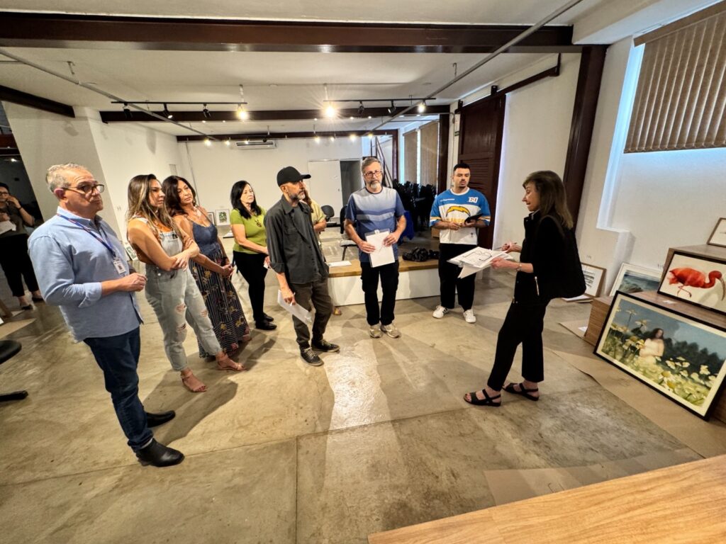 A imagem mostra um grupo de pessoas em um ambiente interno, possivelmente uma galeria de arte ou um espaço de exposição. No centro da cena, uma mulher está falando e gesticulando, segurando papéis, enquanto parece explicar algo para o grupo. O público está disposto em semicírculo ao redor dela, ouvindo atentamente. As pessoas estão vestidas de forma casual e demonstram interesse no que está sendo apresentado. O espaço é amplo, com piso de concreto e teto com vigas expostas e iluminação direcionada, típica de galerias. Na parede à direita, há várias obras de arte emolduradas, algumas apoiadas no chão, incluindo uma pintura que parece ser de um flamingo. A iluminação é suave, criando sombras discretas no chão e destacando os participantes e as obras. A atmosfera é de concentração e aprendizado, sugerindo que se trata de uma apresentação ou visita guiada em uma exposição de arte. As cores predominantes são neutras, com toques de cores vivas nas roupas das pessoas e nas pinturas. A imagem transmite um momento de interação cultural e troca de conhecimento.