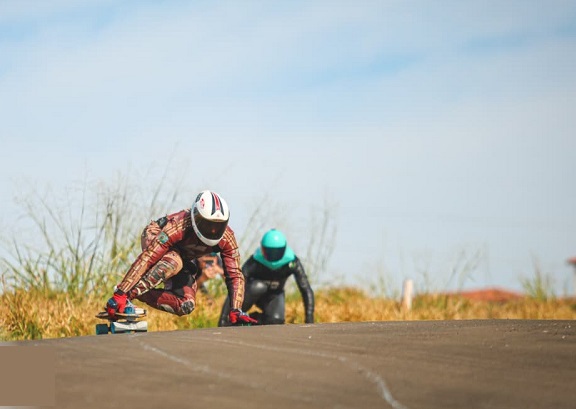 A imagem mostra dois praticantes de longboard descendo uma estrada asfaltada em alta velocidade. O foco principal está no corredor à frente, que está em uma posição aerodinâmica, com o corpo abaixado e uma das mãos tocando o chão para ajudar no equilíbrio. Ele veste um macacão de couro com detalhes em vermelho, preto e dourado, além de um capacete integral branco com detalhes vermelhos. O segundo corredor aparece logo atrás, usando um macacão preto e um capacete integral azul-turquesa. A estrada é cercada por vegetação seca, com gramíneas amareladas e plantas altas nas laterais. O céu está claro, sugerindo um dia ensolarado. A perspectiva da foto é baixa, o que enfatiza a sensação de velocidade e a ação da descida.