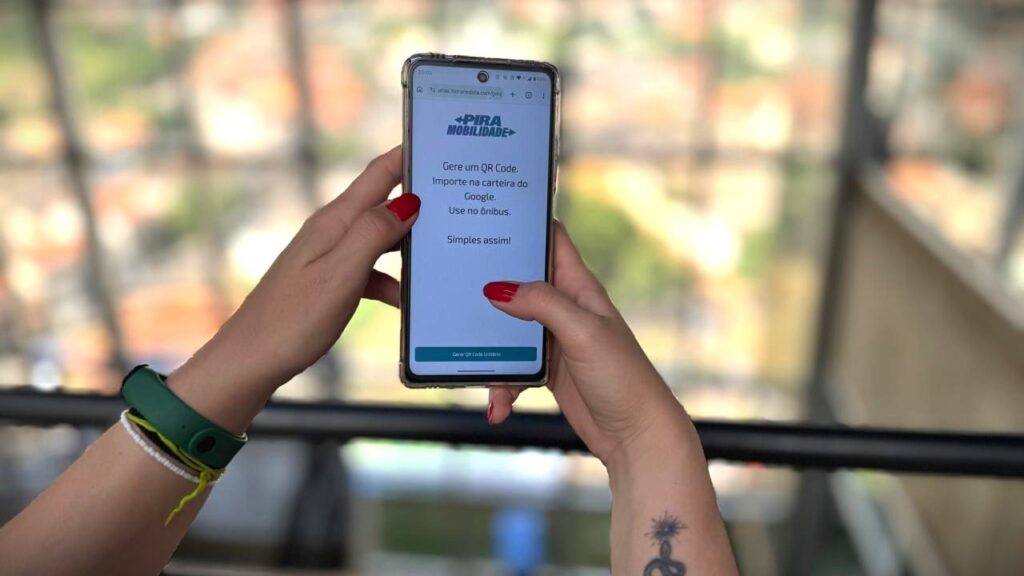 A imagem mostra as mãos de uma pessoa segurando um smartphone na vertical. As unhas estão pintadas de vermelho e no pulso esquerdo a pessoa usa uma pulseira verde de atividade física e uma pulseira branca de corda. No pulso direito há uma pequena tatuagem visível. Na tela do celular, vê-se um aplicativo em português chamado "PIRA MOBILIDADE". O texto na tela orienta o usuário a gerar um código QR para importar na carteira do Google (Google Wallet) e usar no ônibus, com a mensagem "Simples assim!". Há um botão destacado escrito "Gerar QR Code Unitário". O fundo da imagem está desfocado, sugerindo um ambiente externo, possivelmente uma varanda com vista para uma área verde e edificações ao longe. A luz é natural e suave, indicando que a foto foi tirada durante o dia.