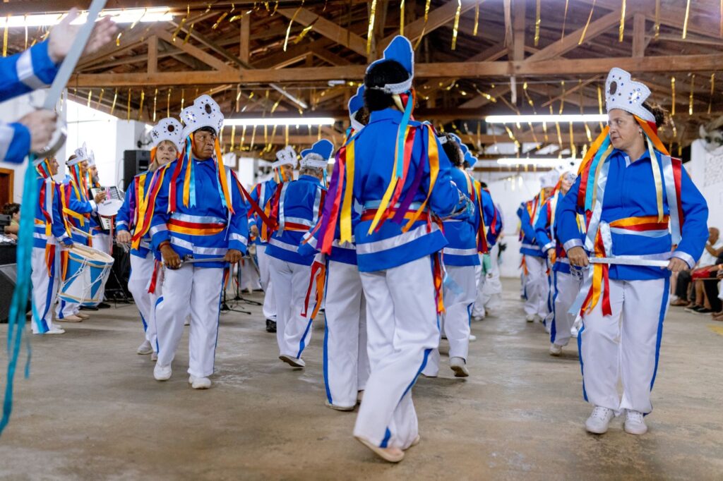 A imagem mostra um grupo de pessoas participando de uma apresentação de dança em um ambiente interno decorado para uma celebração. Os dançarinos estão vestidos com uniformes azuis vibrantes, adornados com fitas coloridas em tons de vermelho e amarelo, que dão um toque festivo à cena. Eles estão organizados em círculo ou semi-círculo, em movimento, transmitindo energia e alegria. O ambiente é iluminado de forma quente, com detalhes que sugerem uma festa tradicional, como a decoração no teto e as estruturas de madeira visíveis. Ao fundo, há equipamentos de som, indicando que se trata de um evento com apresentação formal. A cena expressa um momento cultural e comunitário, com destaque para a coordenação e o entusiasmo dos participantes.
