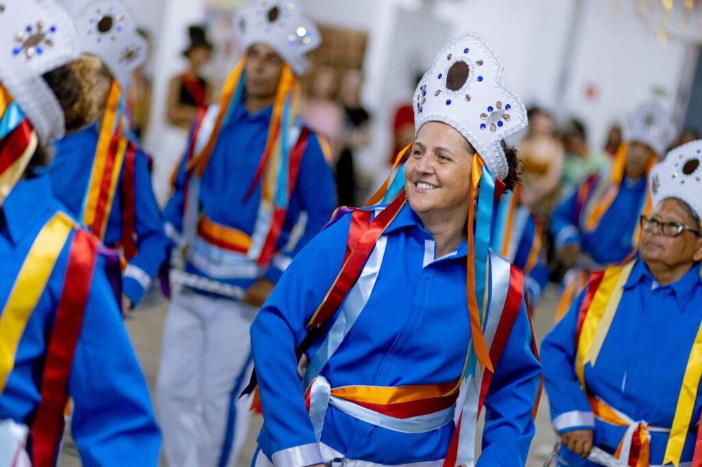 A imagem mostra uma cena vibrante de uma apresentação cultural, provavelmente um desfile ou festival. No centro da imagem, há uma mulher sorridente vestida com uma camisa azul brilhante e mangas compridas, adornada com fitas coloridas em vermelho, laranja, amarelo e azul, que estão amarradas em sua cintura e caem pelos ombros. Ela usa uma coroa branca em forma de estrela, decorada com pequenos brilhos e pedras azuis, com fitas semelhantes penduradas. Ao seu redor, outras pessoas também vestem roupas azuis e trajes festivos, participando da dança ou apresentação. O fundo está desfocado, mas é possível perceber mais participantes e espectadores, sugerindo um evento público movimentado. A imagem transmite uma atmosfera alegre e cheia de energia, destacando o orgulho cultural e a união do grupo.