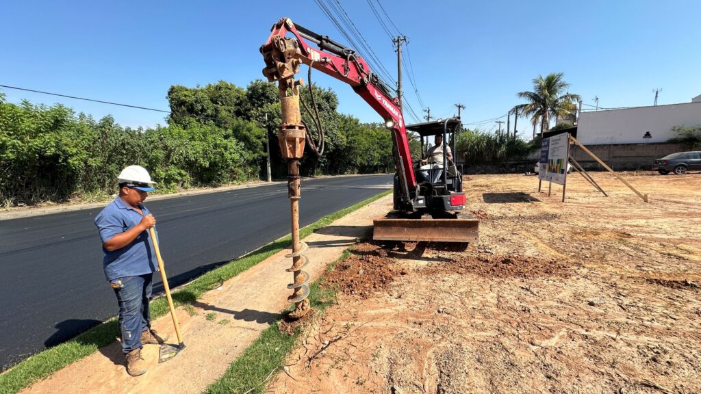 A imagem mostra um canteiro de obras ao ar livre em um dia ensolarado. No centro, há uma mini escavadeira vermelha com um acessório tipo hélice (broca) acoplado, que está sendo usada para perfurar o solo. À esquerda, um homem está parado sobre o asfalto, vestindo uma camisa azul, calça jeans, botas e um capacete branco de segurança. Ele segura uma ferramenta longa, possivelmente uma pá ou enxada, e parece estar observando ou aguardando para trabalhar. O chão ao redor da escavadeira é de terra revolvida, contrastando com a pista asfaltada ao lado. Ao fundo, há árvores verdes e vegetação, além de postes com fios elétricos e um carro estacionado. Também é visível uma placa com texto e imagens, onde se lê "Água tem benefícios" e "Governo Federal", indicando que possivelmente a obra está relacionada a algum projeto público voltado para a água ou saneamento. A iluminação é forte, com sombras marcadas, indicando que a foto foi tirada durante o dia.