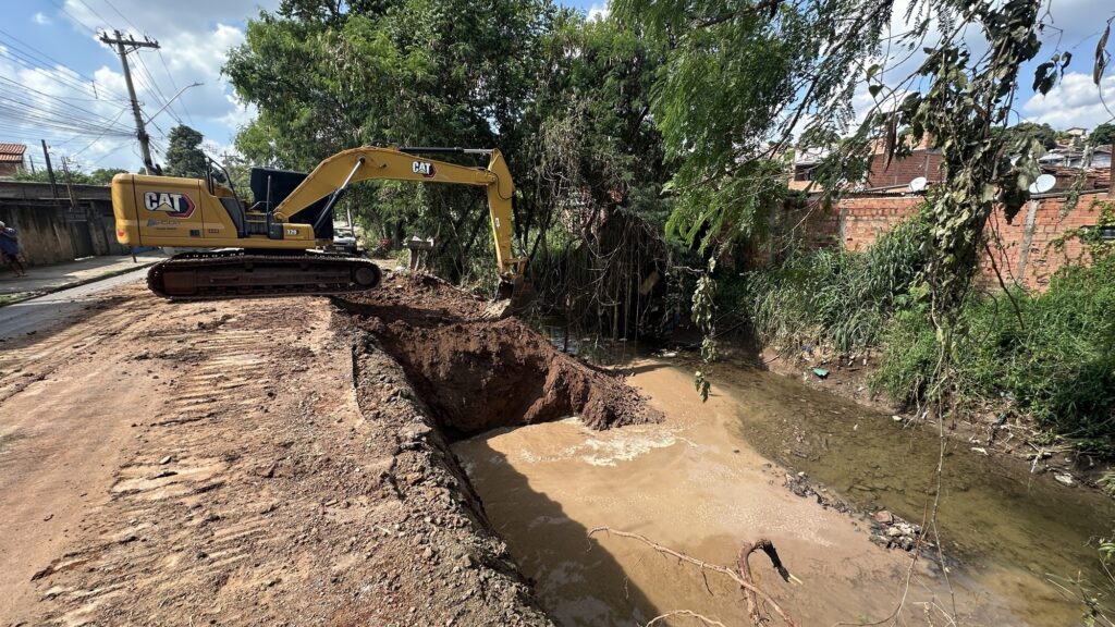 A imagem mostra um canteiro de obras onde uma escavadeira amarela da marca CAT, modelo 320, está escavando a margem de um canal estreito e lamacento. A escavadeira está posicionada sobre uma estrada de terra ao lado do canal, com a concha ativa cavando o solo e criando um grande buraco, deslocando muita terra. O canal contém água turva e marrom, e suas margens são cobertas por vegetação e detritos. Ao redor, há árvores verdes e uma vegetação densa, indicando um ambiente semiurbano. Ao fundo, é possível ver algumas casas de tijolos com telhados e antenas parabólicas, além de postes e fios de energia, o que sugere que a área é habitada. A cena ocorre durante o dia, com luz natural iluminando o local. Um homem está parado na estrada de terra, observando a obra. A escavadeira tem inscrições visíveis, como a marca "CAT", o modelo "320" e um contato com o número "-3435-9000". No geral, a imagem retrata um trabalho de escavação em andamento, possivelmente para manutenção ou limpeza do canal, mostrando a intervenção humana e o uso de maquinário pesado para alterar a paisagem natural e urbana.