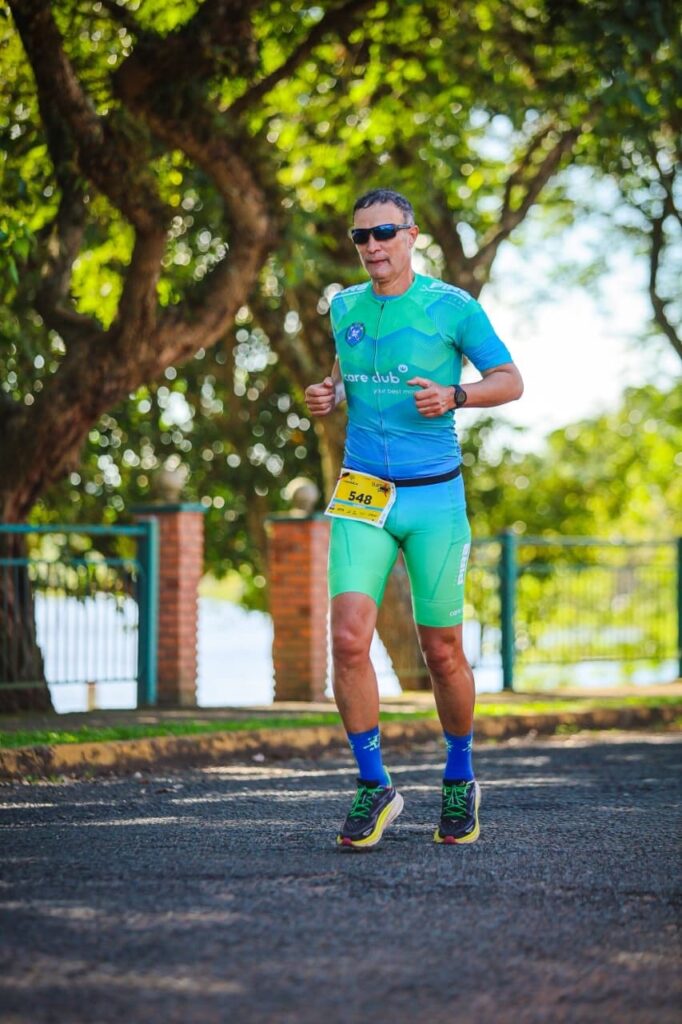 A imagem mostra um homem de meia-idade correndo ao ar livre em uma estrada pavimentada. Ele veste uma camiseta esportiva azul e verde, com um logotipo circular no peito esquerdo, e shorts verdes fluorescentes. Ele usa uma faixa de número de corrida "548" presa ao shorts, além de meias compressivas azuis e tênis pretos com detalhes amarelos e cadarços verdes. O homem está usando óculos escuros e parece concentrado enquanto corre. Ao fundo, há árvores verdes que criam um ambiente natural e sombreado, além de uma cerca de tijolos e um corpo d’água, sugerindo que ele está em um parque ou área de lazer. A luz do sol ilumina a cena, indicando que é durante o dia.
