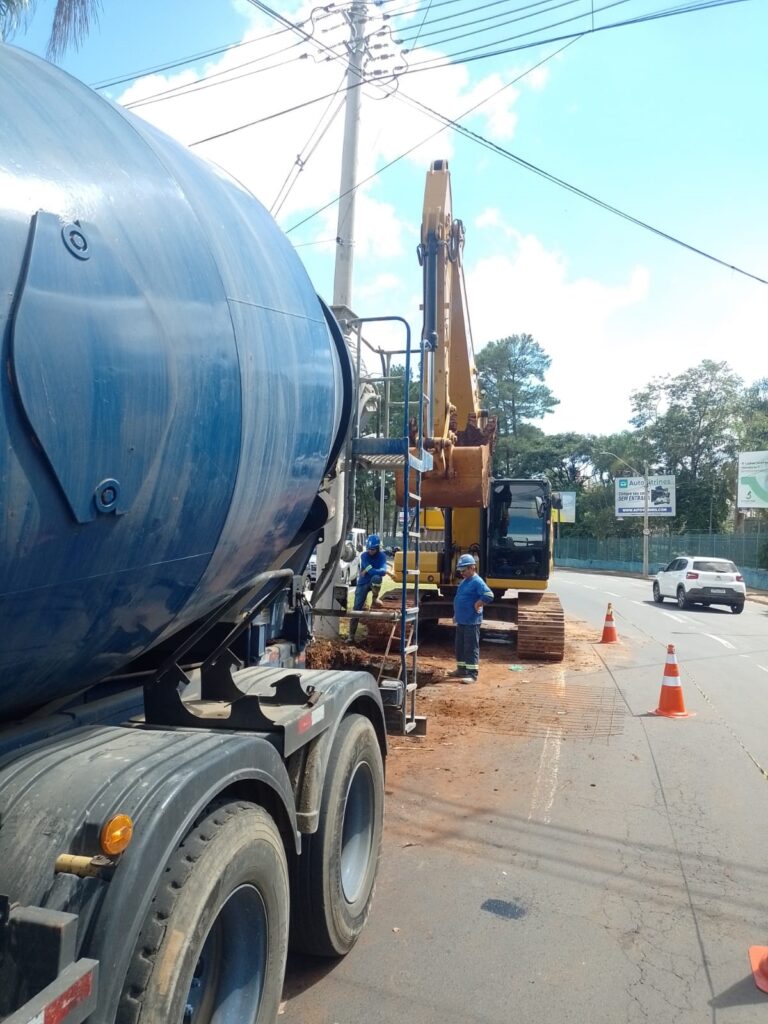 A imagem mostra uma cena de construção na beira de uma estrada em um dia claro e ensolarado. No primeiro plano à esquerda, há um caminhão betoneira azul estacionado. Mais ao centro, um trator escavadeira amarelo está em operação, com a caçamba levantada. Próximo à escavadeira, há dois operários vestindo uniformes azuis e capacetes de segurança, um deles está em pé e o outro sentado na lateral da máquina. À direita, três cones de sinalização laranja delimitam a área de trabalho, enquanto ao fundo, um carro branco segue pela via. Ao fundo, há árvores verdes e um outdoor com a inscrição "Autovitrines", que também exibe o site "WWW.AUTOVITRINES.COM" e a frase "Compre seu carro SEM ENTRADA". A cena transmite uma atividade típica de manutenção ou construção viária em um ambiente urbano ou suburbano.
