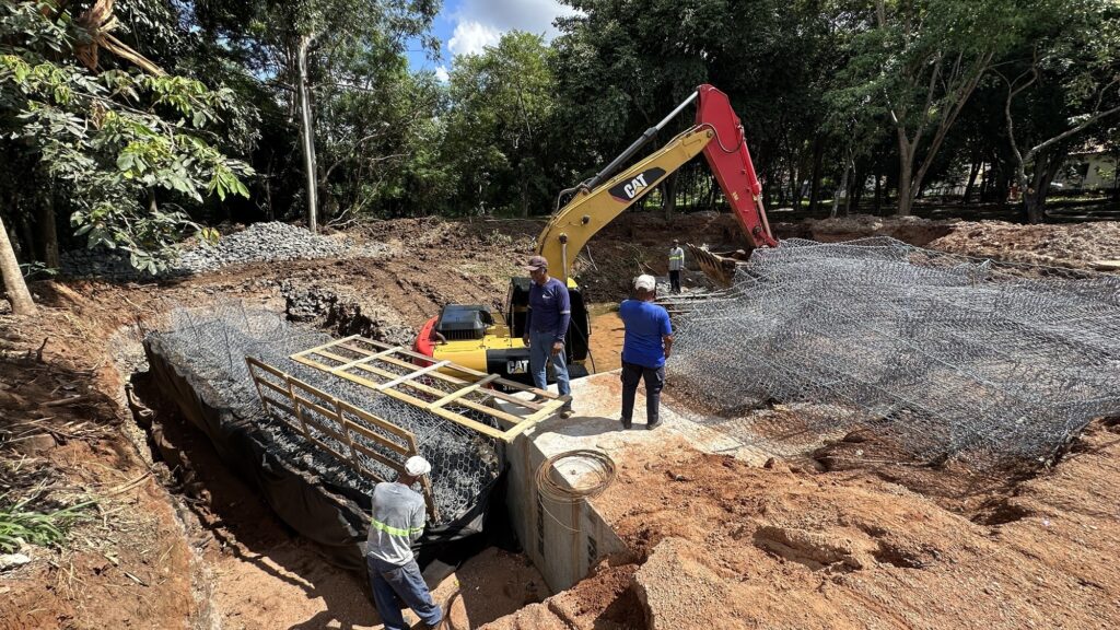 A imagem mostra uma cena de construção ao ar livre, onde uma escavadeira amarela da marca CAT (modelo 310) está operando em uma área de terra e lama, possivelmente um buraco ou fundação. A escavadeira está com a lança estendida, aparentemente movimentando ou colocando material no local. Três homens estão presentes na cena: dois deles estão em primeiro plano observando o trabalho da máquina, um veste uma camisa azul clara e boné branco, o outro está de camisa azul escura e calça jeans; um terceiro homem, com camisa azul clara, está mais ao fundo, próximo à escavadeira. Ao redor do local há vegetação verde e árvores, indicando que a obra está em uma área aberta, cercada pela natureza. O chão está coberto por terra, pedras e estruturas de malha metálica (provavelmente gabiões) utilizadas para contenção ou reforço. Também há tábuas de madeira no local, possivelmente para suporte temporário ou para construção de formas. A iluminação natural do dia destaca a atividade da obra e o ambiente ao redor.