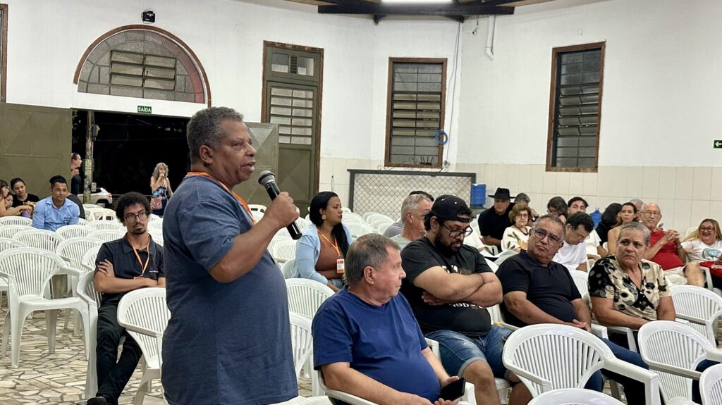 A imagem mostra um homem de meia-idade, com cabelos grisalhos, vestido com uma camiseta azul, falando ao microfone em um ambiente interno que parece ser uma sala de reunião ou auditório comunitário. Ele está em pé, à esquerda da imagem, enquanto várias pessoas estão sentadas em cadeiras plásticas brancas, prestando atenção nele. O público é composto por homens e mulheres de diferentes idades, alguns observando atentamente, outros parecendo mais descontraídos ou distraídos. O ambiente é iluminado artificialmente, com paredes brancas, janelas com venezianas de madeira e um piso de pedras. Ao fundo, há uma porta com uma placa verde escrita "SAÍDA", indicando que o local provavelmente fica em um país de língua portuguesa, possivelmente Brasil. A cena sugere uma reunião comunitária, palestra ou debate, com um clima informal e participativo.