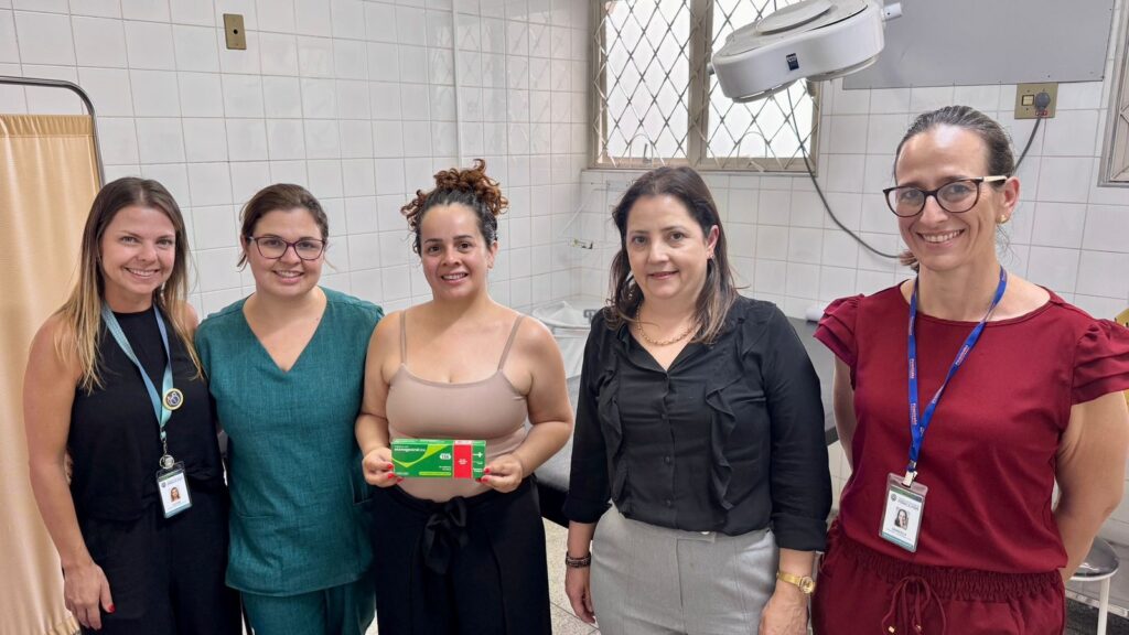 A imagem mostra cinco mulheres posando juntas em um ambiente que parece ser uma clínica ou consultório médico, com paredes claras e piso de azulejo. Elas estão sorrindo e parecem estar felizes. Duas delas vestem roupas casuais, uma está usando jaleco branco, outra usa um uniforme de enfermeira ou técnico de saúde, e a última está com uma roupa social. A mulher no centro segura um cartão ou uma placa com cores vermelha e verde. Ao fundo, é possível ver equipamentos médicos e uma janela que deixa entrar luz natural, iluminando bem o ambiente. A atmosfera é profissional e acolhedora, transmitindo uma sensação de colaboração e amizade entre as mulheres.