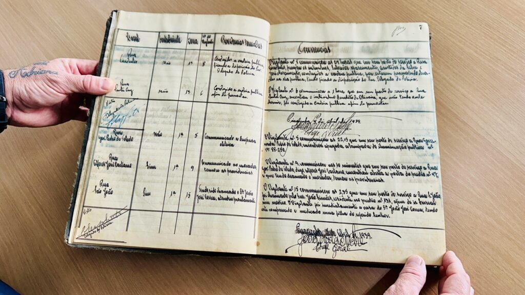 A imagem mostra um livro antigo aberto, possivelmente um registro oficial ou um diário de ocorrências. As páginas são amareladas pelo tempo e contêm textos manuscritos em português, escritos com tinta preta. No lado esquerdo, há uma tabela com colunas para nomes, endereços e outras informações, enquanto a página da direita está intitulada "Ocorrências" e contém relatos detalhados de eventos ou incidentes, também escritos à mão. A mão de uma pessoa com tatuagens está segurando a página esquerda, indicando o manuseio cuidadoso do documento. O texto manuscrito é denso e parece registrar informações oficiais, possivelmente relacionadas a atendimentos policiais ou administrativos, com menções a nomes, datas e ações tomadas. O ambiente parece ser interno, com iluminação natural suave que destaca a textura envelhecida do papel e o estilo da caligrafia antiga. A superfície onde o livro está apoiado parece ser de madeira, complementando o aspecto histórico da cena. A imagem transmite uma sensação de importância documental e preservação histórica.