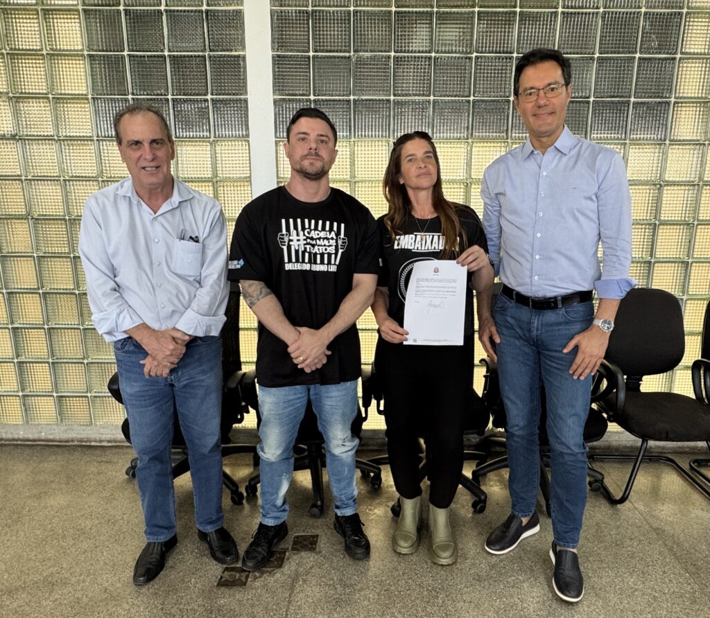 Na imagem aparecem três homens e uma mulher. No lado direito aparece um senhor de camisa clara, calça jeans clara e sapatos pretos e do lado esquerdo da mulher que está com roupa preta, segurando uma folha, está um homem de óculos, camisa azul clara e calça jeans escura. Estão em uma sala cujo vidros aparecem atrás.