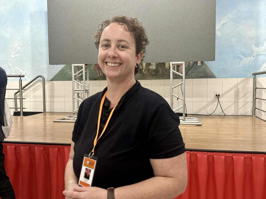 A imagem mostra uma mulher sorridente com cabelo curto e encaracolado, vestindo uma camisa polo preta e um crachá laranja pendurado no pescoço. Ela está em um ambiente interno, provavelmente um salão de eventos ou uma escola, diante de um palco de madeira com uma tela digital grande em cinza, sustentada por estruturas metálicas. Ao fundo, há uma parede decorada com um mural que representa um céu azul com nuvens e pássaros brancos. A iluminação é quente, criando um ambiente acolhedor. A mulher está em foco, em posição central na foto, transmitindo uma sensação de simpatia e cordialidade.
