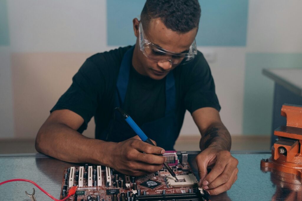 A imagem mostra um jovem concentrado realizando um trabalho técnico em uma placa-mãe de computador. Ele usa óculos de proteção transparentes, uma camiseta preta e um avental azul, indicando que está em um ambiente de trabalho ou laboratório. O jovem está segurando um ferro de solda com uma das mãos, enquanto a outra mão apoia a placa-mãe sobre uma bancada metálica. Ao lado da bancada, há uma morsa laranja, sugerindo que o local é equipado para trabalhos manuais e eletrônicos. A placa-mãe é da marca ASUS, modelo NF7-S, conforme indicado pelas inscrições visíveis na peça. O ambiente parece ser um laboratório ou oficina, com paredes claras ao fundo e iluminação natural que destaca o trabalho detalhado que está sendo feito. A imagem transmite uma atmosfera de concentração, habilidade técnica e cuidado no manuseio dos componentes eletrônicos.