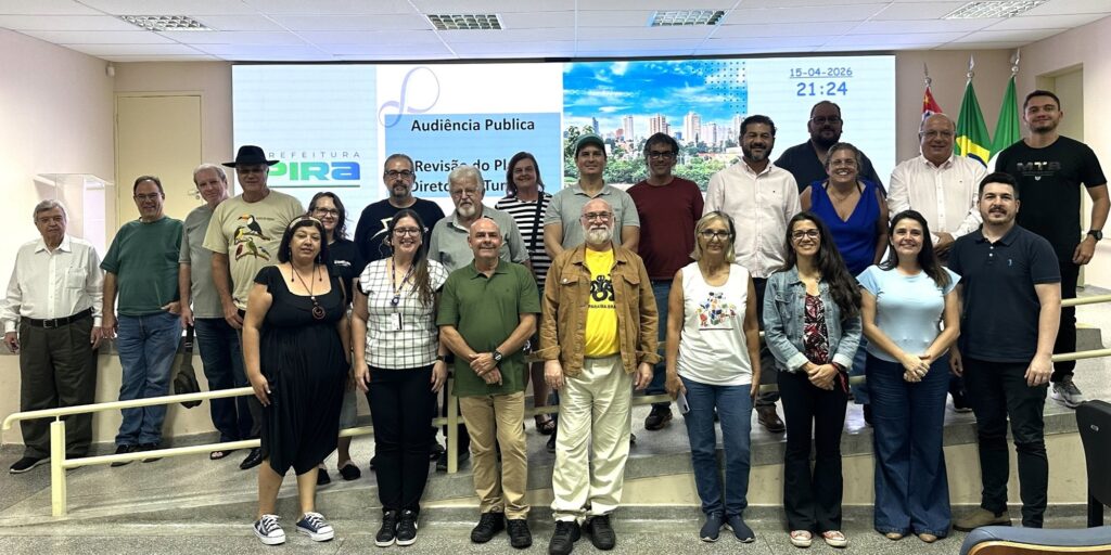 A imagem mostra um grupo de 21 pessoas reunidas para uma foto em um ambiente interno, possivelmente uma sala de reuniões ou auditório. Ao fundo, há uma grande tela projetando informações sobre o evento: "Audiência Pública - Revisão do Plano Diretor - Turismo", datada de 15 de abril de 2026, às 21h24. Também aparece o logo da Prefeitura de Pira, indicando que o evento é oficial e ligado à administração municipal. As pessoas estão organizadas em três níveis: algumas sentadas no chão, outras agachadas e o restante em pé atrás. A maioria está sorrindo ou com expressões neutras, vestindo roupas casuais e semi-formais. No canto direito, é possível ver algumas bandeiras, provavelmente a bandeira do Brasil e outras bandeiras estaduais ou municipais. O ambiente é iluminado artificialmente, com uma luz que destaca o grupo e a tela ao fundo, criando uma atmosfera formal e de colaboração coletiva. A foto parece ter sido tirada para registrar a participação dos presentes na audiência pública relacionada ao planejamento urbano e turismo da cidade.