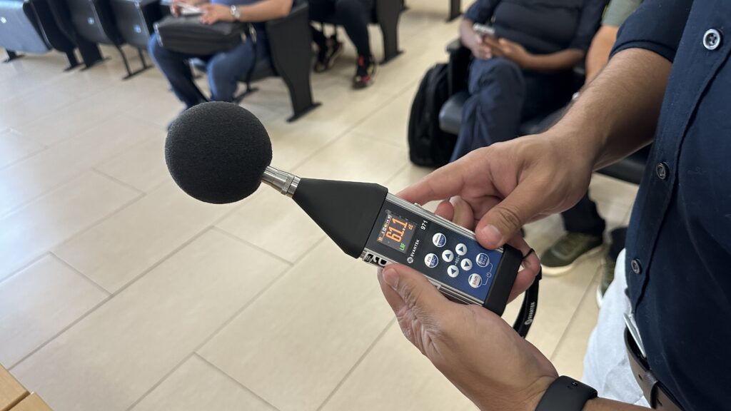 A imagem mostra as mãos de uma pessoa segurando um medidor de nível sonoro da marca Svantek, modelo 971. O aparelho está ligado e exibe uma leitura de 61,1 decibéis (dB) na tela digital, com o indicador "LAF", que se refere a um tipo de ponderação e tempo de resposta do som. O medidor possui um microfone com uma cobertura esférica preta, usada para proteger contra o vento e ruídos externos. Ao fundo, desfocado, há um ambiente interno com pessoas sentadas em cadeiras pretas, sugerindo que a medição está sendo feita em um local público, possivelmente uma sala de espera ou auditório. A pessoa que segura o aparelho veste uma camisa azul escura e está posicionada de forma que o medidor está em destaque na imagem, em foco, enquanto o fundo está desfocado para dar ênfase ao equipamento e à leitura do nível sonoro.