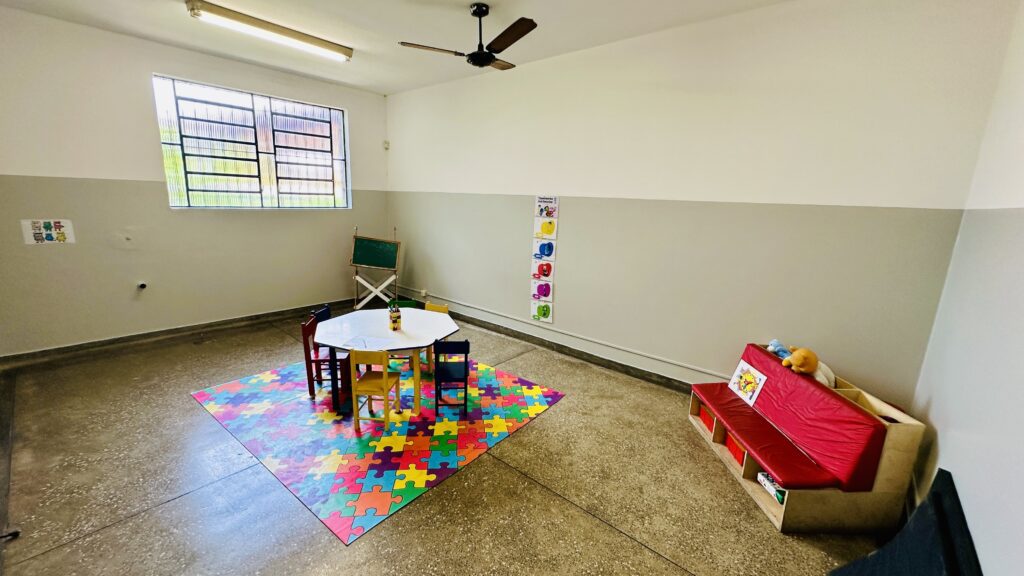 A imagem mostra uma sala de aula ou área de recreação para crianças pequenas. No centro, há uma mesa branca em formato hexagonal, rodeada por quatro cadeiras pequenas, adequadas para crianças. A mesa está sobre um tapete colorido com padrão de peças de quebra-cabeça, que cobre boa parte do chão de cimento. À direita do tapete, há um banco vermelho com estantes de madeira embutidas embaixo, onde estão guardados livros e brinquedos. As paredes são pintadas em duas cores: a parte superior é creme claro e a parte inferior é cinza claro, separadas por uma linha horizontal. Uma janela grande com grades metálicas está à esquerda, permitindo a entrada de luz natural. Acima da mesa, há um ventilador de teto. Perto da janela, há cartazes coloridos fixados na parede. Atrás da mesa, há um pequeno quadro-negro verde em um cavalete. A iluminação da sala combina luz natural da janela com luz artificial de uma lâmpada fluorescente no teto. O ambiente transmite uma sensação de organização, limpeza e é claramente pensado para o conforto e aprendizado das crianças.
