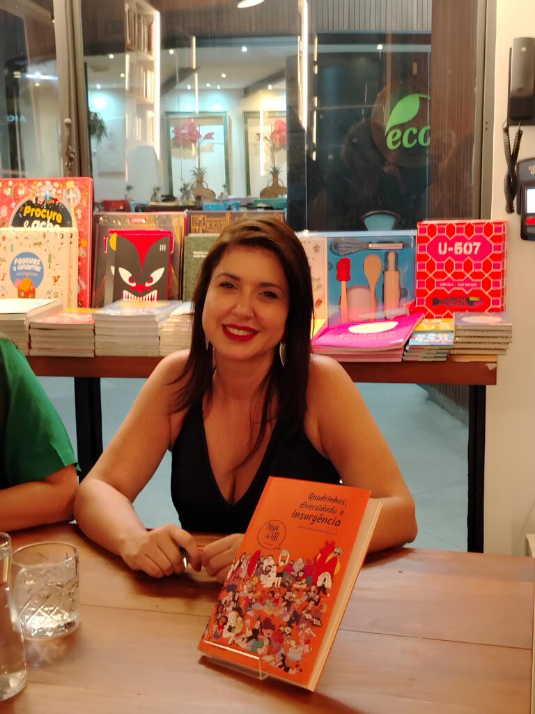A imagem mostra uma mulher sorridente, de cabelos escuros, sentada à mesa de um ambiente que parece ser uma livraria ou cafeteria. Ela veste uma blusa preta de decote e está diante de um livro laranja com uma ilustração colorida na capa. Na mesa, há também um copo de vidro com água. Ao fundo, há prateleiras repletas de livros, incluindo títulos infantis como "Procure e ache" e "Procure e encontre". Também é possível ver alguns objetos coloridos, possivelmente brinquedos ou materiais educativos. A iluminação é quente e destaca o rosto da mulher e os itens sobre a mesa, criando um ambiente acolhedor e convidativo. No fundo, há um logotipo verde com a palavra "eco", que sugere um local com preocupação ambiental. A composição da foto é feita em um ângulo levemente superior, focando na mulher e no livro à sua frente.