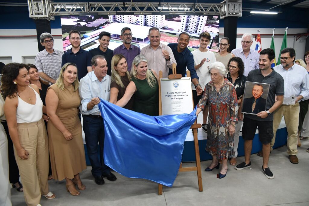 A imagem mostra um evento comemorativo realizado em um ambiente interno, provavelmente uma cerimônia municipal ou comunitária. Um grupo de cerca de 20 pessoas está reunido em torno de uma placa ou cartaz grande, que parece ter sido recentemente revelado, indicando o lançamento ou inauguração de um projeto ou iniciativa da prefeitura. As pessoas estão vestidas de forma mista, com alguns usando roupas formais e outros mais casuais, transmitindo um clima de celebração e formalidade ao mesmo tempo. No fundo, há uma tela digital exibindo imagens ou plantas arquitetônicas, possivelmente relacionadas ao projeto que está sendo apresentado. Também são visíveis duas bandeiras, o que reforça o caráter oficial do evento. No centro da imagem, uma mulher está no ato de revelar a placa, enquanto os demais participantes observam, sorriem e interagem entre si. Um homem segura uma moldura com uma foto ou imagem importante, sugerindo que pode haver uma homenagem ou reconhecimento durante a cerimônia. A iluminação é clara e destaca bem as pessoas e os elementos principais da cena, indicando que o momento é importante e está sendo registrado com atenção. No geral, a imagem transmite uma sensação de orgulho comunitário e envolvimento coletivo em um projeto público.