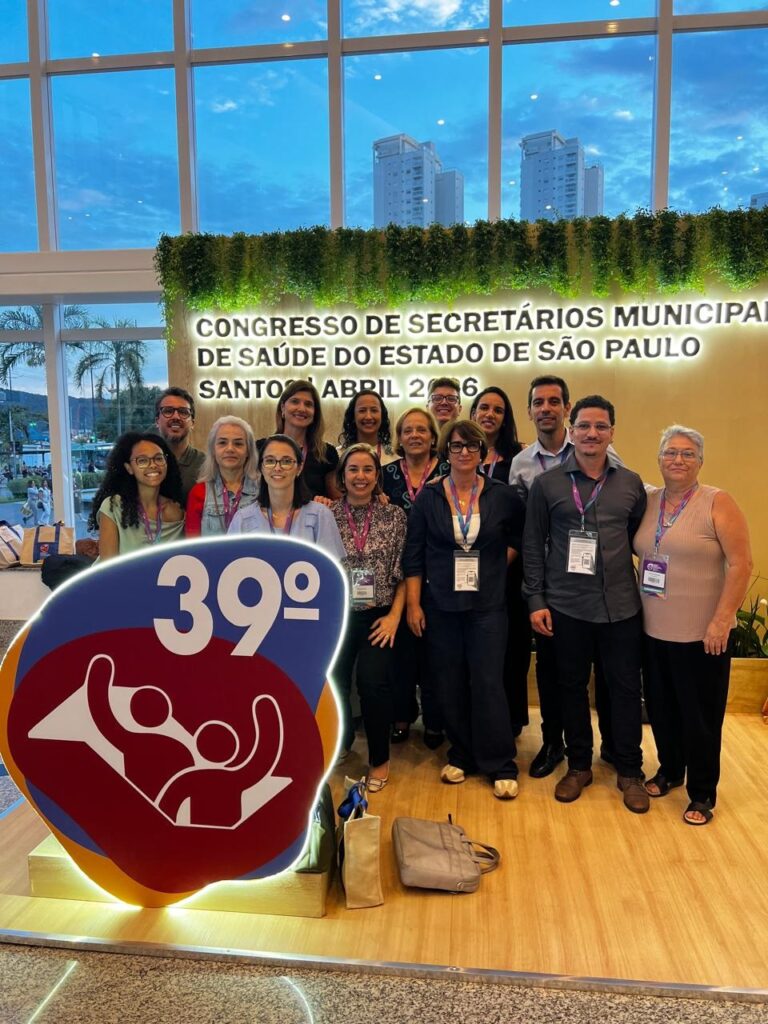 A imagem mostra um grupo de cerca de 15 pessoas posando para uma foto em um ambiente interno, possivelmente um centro de convenções ou um salão de conferências. O grupo é composto por homens e mulheres, todos vestidos de forma casual e profissional. Ao fundo, há um grande banner com o texto "CONGRESSO DE SECRETÁRIOS MUNICIPAL DE SAÚDE DO ESTADO DE SÃO PAULO" e a indicação "39°", sugerindo que se trata da 39ª edição desse congresso. Também aparece a palavra "SANTO ABRIL 2 6", possivelmente referindo-se à data ou local do evento. À esquerda da imagem, há um logo colorido com o número "39°" em destaque e uma figura estilizada de duas pessoas. O ambiente é moderno, com uma parede verde decorada com plantas, janelas grandes que permitem a entrada de luz natural e uma iluminação artificial que clareia bem o espaço. No chão, perto do grupo, há algumas bolsas ou sacolas. A expressão das pessoas é sorridente e amigável, indicando um clima positivo e de confraternização durante o evento.
