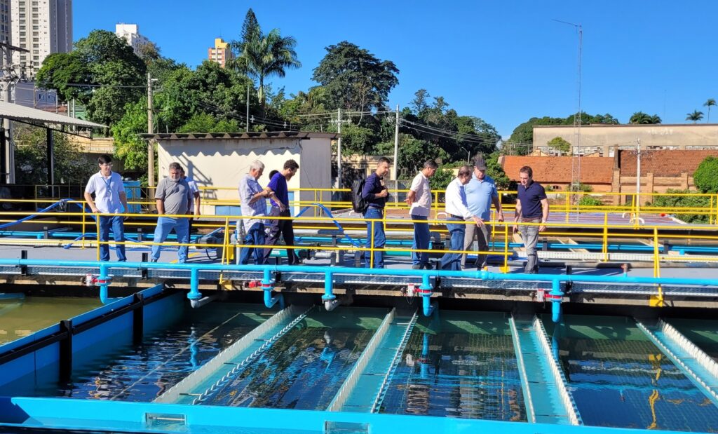 A imagem mostra um grupo de dez homens posando para uma foto em uma estrutura industrial ao ar livre, que parece ser uma estação de tratamento de água. Eles estão em uma plataforma de concreto com corrimões amarelos, acima de tanques ou piscinas de tratamento de água visíveis na parte inferior da imagem. Ao fundo, há vegetação densa e árvores, indicando que o local está cercado por área verde. Também é possível ver postes de energia elétrica e uma construção alta ao longe. Os homens estão vestidos de forma casual e semi-formal, com camisas e calças, e parecem estar atentos à câmera, alguns sorrindo. A iluminação indica um dia claro e ensolarado, com o céu azul sem nuvens. Na parte de trás da plataforma, há vários contêineres amarelos, um dos quais tem a inscrição "COTEVIA". A composição da foto é equilibrada, com o grupo centralizado e o ambiente industrial e natural formando o cenário ao redor. A imagem sugere uma visita técnica ou inspeção no local de tratamento de água.