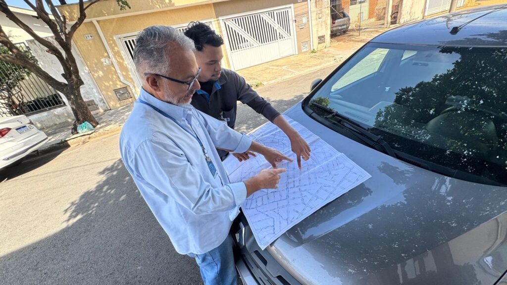 A imagem mostra dois homens em uma rua residencial, ambos concentrados em um grande mapa ou planta que está apoiado no capô de um carro branco. O homem à frente, de cabelos grisalhos e barba, veste uma camisa azul clara e usa óculos e um crachá pendurado no pescoço. Ele está apontando para o mapa com a mão esquerda. O segundo homem, mais jovem, veste uma camiseta cinza escura e também está apontando para o mapa, observando atentamente. Ao fundo, vemos casas com muros brancos e portões, além de uma árvore com poucas folhas. A cena acontece durante o dia, com luz natural e sombras indicativas de sol. A imagem sugere que os homens estão envolvidos em uma atividade de planejamento, análise ou orientação usando o mapa.