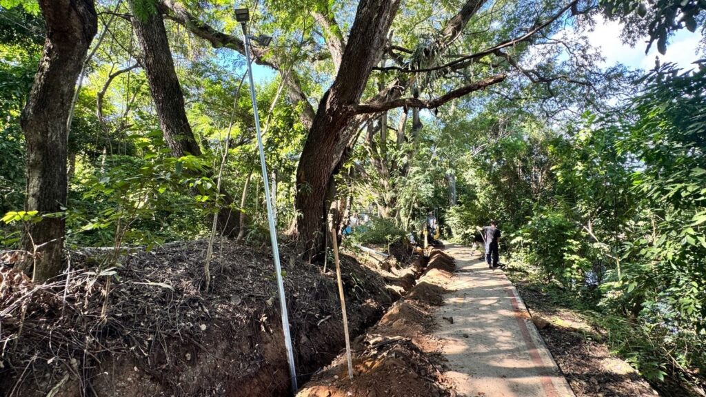 A imagem mostra uma trilha pavimentada, cercada por árvores grandes e vegetação densa e verde. Ao lado da trilha, há uma vala aberta, onde parece estar sendo feita uma obra, possivelmente instalação ou manutenção de postes, já que há um poste metálico instalado dentro da vala. Também é possível ver estacas de madeira ao redor da vala, provavelmente para demarcação. Ao fundo, há uma escavadeira amarela, indicando que o trabalho é recente e envolve maquinário pesado. Duas pessoas caminham pela trilha, afastando-se da câmera. A cena é iluminada pela luz do dia, com sombras das árvores cobrindo parte do caminho, criando um ambiente natural e tranquilo, apesar da obra em andamento.