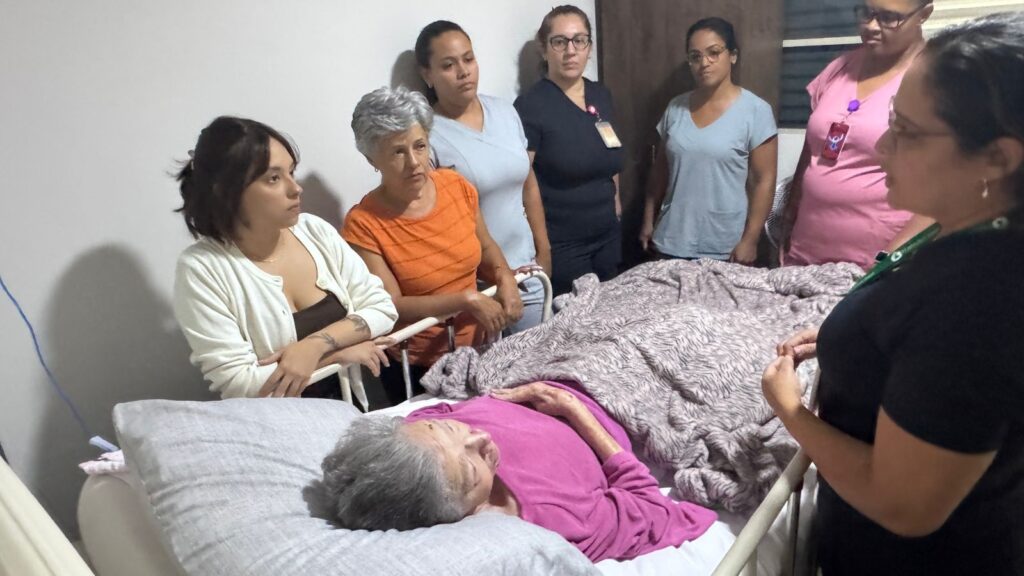 A imagem mostra um grupo de seis mulheres reunidas em torno de uma mulher idosa que está deitada em uma cama hospitalar. A mulher idosa, que parece frágil, está deitada de costas, apoiada por travesseiros e coberta por um cobertor com estampa. As outras mulheres estão em pé ao redor da cama, olhando para ela com expressões de cuidado e atenção, sugerindo um ambiente de apoio e preocupação. Algumas delas usam roupas que lembram uniformes hospitalares, como jalecos ou scrubs, enquanto outras vestem roupas casuais. A iluminação é suave e a cena transmite um sentimento de acolhimento e solidariedade, possivelmente em um ambiente hospitalar ou de cuidado de saúde.