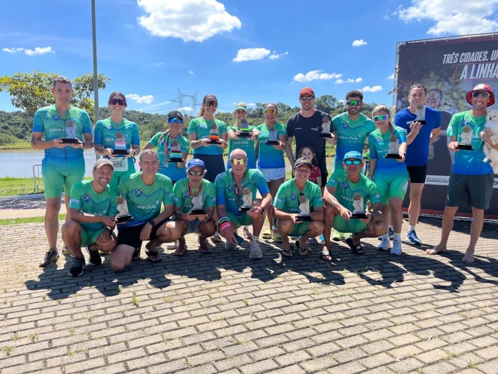 A imagem mostra um grupo de cerca de 19 pessoas, todas vestindo roupas esportivas, predominantemente em tons de azul e verde, com o logo "care club" estampado nas camisetas. A maioria está segurando troféus, indicando que participaram de uma competição esportiva e conquistaram premiações. O grupo está posicionado ao ar livre, em uma área pavimentada, com um lago ou reservatório e vegetação ao fundo. O céu está claro com poucas nuvens, sugerindo um dia ensolarado. As pessoas parecem felizes e orgulhosas, algumas estão em pé enquanto outras estão agachadas ou sentadas no chão, formando duas fileiras. Um cachorro branco pequeno também aparece junto ao grupo. Ao fundo, há uma faixa com texto em português, parcialmente visível, mencionando "TRÊS CIDADES" e "A LINHA", sugerindo o nome ou tema do evento. No geral, a imagem transmite um momento de celebração e conquista após uma atividade esportiva coletiva, possivelmente uma corrida ou prova de resistência, realizada em um ambiente natural e agradável.