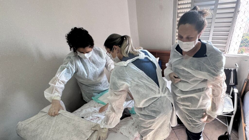 A imagem mostra três pessoas vestidas com equipamentos de proteção individual (EPI), incluindo aventais descartáveis, máscaras e luvas, provavelmente profissionais de saúde ou cuidadores. Elas estão em um ambiente doméstico, possivelmente um quarto, dado que há uma cama com roupa de cama estampada e uma cabeceira de madeira. Uma das pessoas está ajustando o travesseiro ou cobertor da cama, outra está inclinada sobre a cama, aparentemente cuidando de algo ou alguém, e a terceira está em pé, observando ou auxiliando. Ao fundo, é possível ver parcialmente uma cadeira de rodas, o que sugere que podem estar cuidando de uma pessoa com mobilidade reduzida. A iluminação é suave e natural, e o ambiente transmite uma sensação de cuidado e atenção.