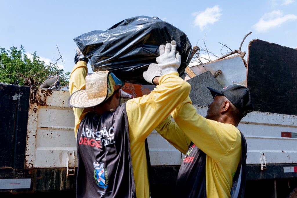 A imagem mostra dois homens colaborando para levantar um grande saco preto de lixo e colocá-lo na caçamba de um caminhão carregado com entulhos. Ambos vestem coletes pretos com inscrições e brasões, além de camisas amarelas de manga longa e luvas brancas. Um deles usa um chapéu de palha, enquanto o outro usa boné preto. O caminhão é branco, com sinais de uso, e está cheio de restos de madeira, metal e outros materiais descartados. Ao fundo, vê-se um céu azul com algumas nuvens e árvores verdes à esquerda. A cena sugere uma ação de limpeza ou coleta de resíduos, em um dia ensolarado. A imagem transmite um senso de cooperação e trabalho em equipe.
