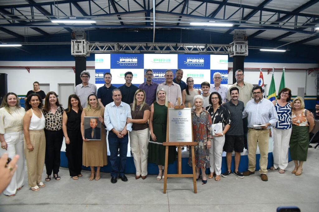 A imagem mostra um grupo de pessoas posando para uma foto em um ambiente interno, possivelmente durante uma cerimônia ou evento oficial. O grupo está disposto em duas fileiras, com algumas pessoas sentadas e outras em pé. No centro da imagem, em destaque, há uma placa em um cavalete de madeira que anuncia a inauguração da "Escola Municipal Professor Humberto de Campos". Ao fundo, há um palco com banners que exibem o nome "Prefeitura de Piracicaba" e o brasão do município, além de duas bandeiras — uma do Brasil e outra do estado de São Paulo. As pessoas na foto estão sorrindo, vestidas de forma formal e semiformal, indicando que se trata de uma ocasião comemorativa ou oficial. A iluminação é clara e uniforme, destacando bem os rostos e a placa. A atmosfera geral da imagem é de celebração e formalidade, sugerindo um momento importante para a comunidade local.