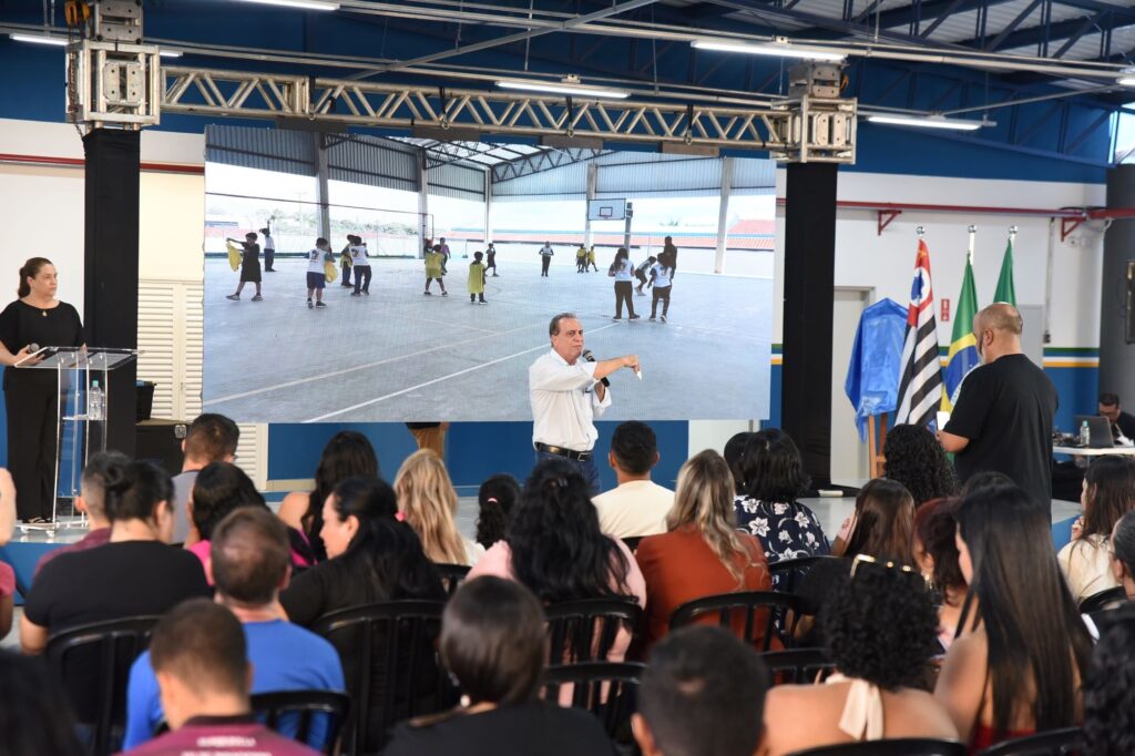 A imagem mostra um evento realizado em um ambiente interno, possivelmente uma escola ou centro comunitário. No centro, há uma grande tela onde é exibido um vídeo de crianças brincando em uma quadra coberta, que parece ser uma quadra de basquete. Em primeiro plano, um homem vestido com camisa branca e calça jeans está falando ao microfone e apontando para a tela, indicando que ele é o palestrante principal. Ao lado dele, uma mulher com roupas pretas está em pé atrás de um púlpito transparente, também segurando um microfone e olhando para a tela. À frente, a audiência está sentada, com as pessoas voltadas para o palco e prestando atenção à apresentação. No fundo, há bandeiras do Brasil e do estado de São Paulo, além de um pano azul decorativo. A iluminação destaca o palco e a tela, enquanto o público está em uma área mais escura. A cena sugere um evento formal relacionado à educação ou projetos comunitários.
