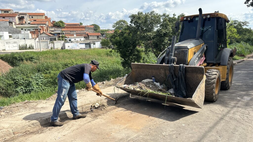 A imagem mostra uma cena de trabalho manual combinada com o uso de maquinário pesado em um ambiente ao ar livre. No primeiro plano, um homem está usando um picareta para quebrar detritos em uma superfície de terra ou asfalto desgastado. Ele veste camiseta preta, calça jeans azul com a inscrição "WMS FACILITIES" na perna, luvas de trabalho e boné. Ao lado dele, à direita, há uma retroescavadeira amarela da marca John Deere, com o balde carregado de lixo e entulho, incluindo sacos plásticos e restos de vegetação. O maquinário tem a inscrição "COTERRA" na cabine. Ao fundo, é possível ver um conjunto de casas com telhados de cerâmica, situadas em um terreno inclinado, rodeadas por bastante vegetação verde. O céu está parcialmente nublado, com nuvens brancas e luz do sol, indicando um dia claro. A cena sugere uma operação de limpeza ou manutenção de estrada, onde o homem quebra o material que provavelmente será recolhido pela retroescavadeira. O contraste entre o ambiente natural e as atividades de limpeza urbana chama atenção para o esforço de manter o local limpo e organizado.