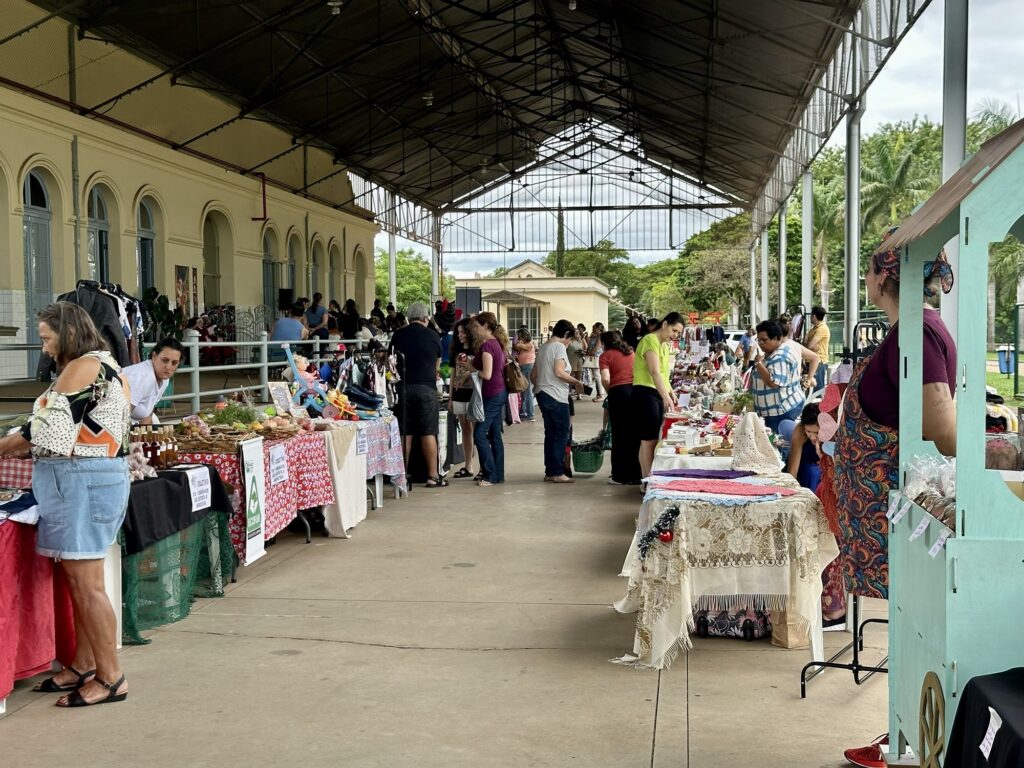 A imagem mostra uma feira ao ar livre sob uma estrutura coberta com um telhado metálico e paredes laterais com janelas arqueadas. No local, há várias barracas organizadas em fileiras, onde são expostos diferentes produtos, como alimentos, artesanatos e roupas. Algumas barracas têm toalhas coloridas e itens dispostos de forma atraente. Diversas pessoas estão circulando pelo espaço, algumas observando os produtos e outras conversando. O ambiente parece movimentado e descontraído, com um clima de comunidade. Ao fundo, pode-se ver árvores e palmeiras, indicando que o local fica em uma região de clima quente ou tropical. A palavra "COLETIVO ECOFAN" aparece em um dos estandes, sugerindo que pode ser um grupo ou projeto que participa da feira, possivelmente relacionado a produtos ecológicos ou sustentáveis. No geral, a cena transmite a sensação de uma feira local, onde moradores e visitantes se reúnem para comprar, vender e socializar em um ambiente aberto e agradável.