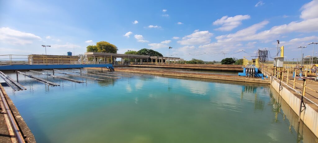 A imagem mostra uma grande estação de tratamento de água ao ar livre em um dia ensolarado com céu azul e algumas nuvens brancas. O foco principal é uma grande bacia retangular cheia de água clara e azul-turquesa. Do lado esquerdo, há várias estruturas paralelas, possivelmente parte de um sistema de filtração ou aeração, feitas de metal com textura canelada, submersas na água. No plano intermediário, aparecem diversas construções de concreto e metal, como passarelas com corrimãos amarelos, prédios de teto plano e tubulações, algumas pintadas de azul. Uma estrutura semelhante a uma ponte azul atravessa a bacia de água à esquerda. Ao fundo, vê-se uma linha de árvores e uma paisagem distante sob o céu claro. A iluminação natural destaca as cores vibrantes da água e do céu, além dos detalhes das estruturas. A composição panorâmica enfatiza a grande escala da estação, com uma perspectiva levemente elevada que permite uma visão ampla do local. Não há pessoas visíveis na imagem, reforçando o ambiente industrial e funcional da estação de tratamento de água.