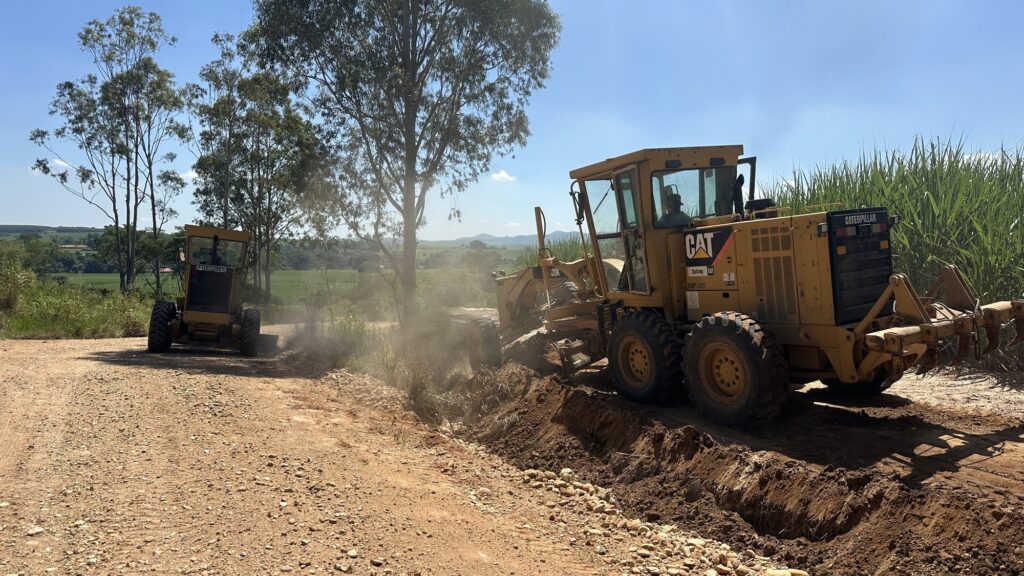 A imagem mostra duas motoniveladoras amarelas da marca Caterpillar trabalhando na manutenção ou construção de uma estrada de terra em uma área rural. A principal motoniveladora está em primeiro plano, posicionada na margem direita da estrada, realizando uma escavação ou nivelamento do solo, levantando poeira ao redor da lâmina. No fundo, outra motoniveladora, menor, está posicionada do lado esquerdo da estrada, provavelmente também em atividade. O cenário é composto por uma estrada de terra cercada por vegetação verde, incluindo árvores altas e uma plantação que parece ser de cana-de-açúcar. Ao fundo, é possível ver colinas suaves sob um céu azul claro, indicando um dia ensolarado. O operador da motoniveladora em primeiro plano está visível dentro da cabine, usando um chapéu. As máquinas exibem as marcas "CAT", "Caterpillar" e "Sotreq", além do modelo "120K VHP-145", evidenciando que são equipamentos pesados modernos da marca reconhecida mundialmente. A imagem está em foco nítido, capturando detalhes tanto nas áreas iluminadas quanto nas sombras, com uma perspectiva ao nível dos olhos do observador.