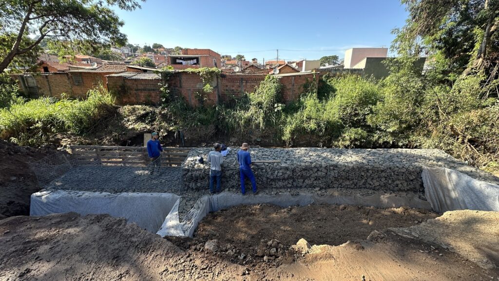 A imagem mostra uma cena de construção ao ar livre, onde trabalhadores estão erguendo uma estrutura de contenção feita com gabiões — caixas de arame preenchidas com pedras. A estrutura está sendo construída em camadas, com pedras sendo cuidadosamente colocadas dentro das gaiolas de arame. Três trabalhadores aparecem na imagem: dois vestem macacões azuis e um está com camiseta cinza e calça jeans. Eles estão ativos no processo, com dois deles posicionados sobre a estrutura de gabiões e um observando próximo. O ambiente é ensolarado, com sombras definidas, indicando que a foto foi tirada durante o dia, provavelmente no final da manhã ou início da tarde. Ao redor da construção, há terra escavada acumulada e vegetação verde, com algumas árvores e plantas que contrastam com o solo e as pedras. No fundo, é possível ver casas com telhados vermelhos, sugerindo que o local é uma área semiurbana ou rural, possivelmente em um terreno inclinado, dado o relevo visível. A estrutura construída parece ter a função de estabilizar o solo, evitar erosão ou servir como base para outra construção, algo comum em obras que utilizam gabiões devido à sua resistência e permeabilidade. A imagem foi capturada de um ângulo levemente elevado, permitindo uma boa visão da profundidade da escavação e do progresso da construção.