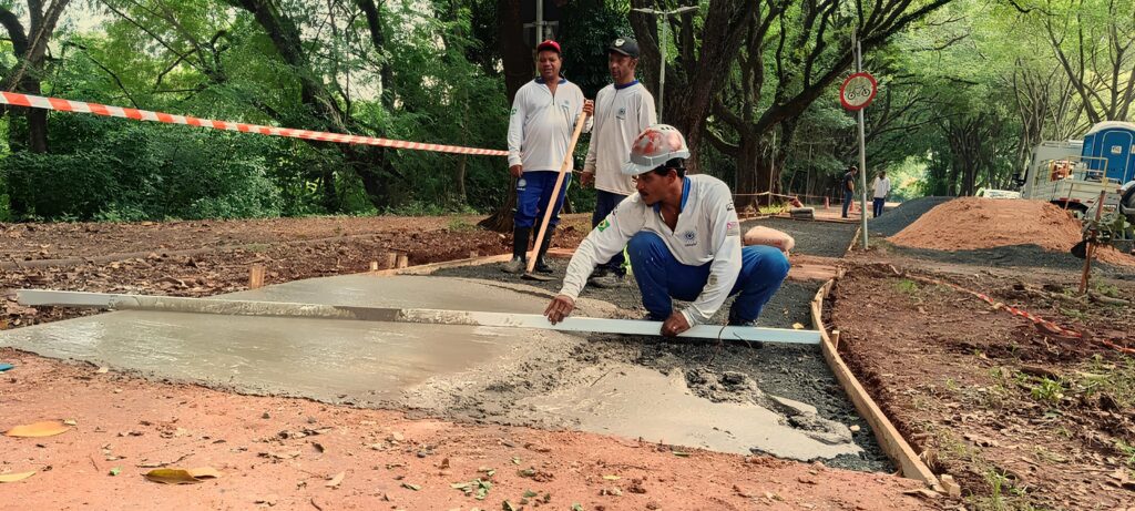 A imagem mostra uma cena de construção ao ar livre, em uma área que parece ser um parque ou espaço verde. No centro da imagem, um trabalhador está agachado, nivelando concreto fresco com uma ferramenta. Ele veste camisa branca, calça azul e capacete, indicando que está em uma obra. O concreto está sendo despejado em um caminho delimitado por formas de madeira, e há uma fita de segurança vermelha e branca sinalizando a área em construção. Ao redor, há terra vermelha e cascalho escuro, além de árvores verdes que cercam o local, sugerindo um ambiente natural. No fundo, aparecem outros trabalhadores, alguns segurando ferramentas, e também é possível ver um caminhão, uma cabine sanitária portátil e uma placa de trânsito com símbolo de bicicleta, indicando que o caminho pode ser para ciclistas. O ambiente está bem iluminado, provavelmente durante o dia, e a cena transmite a ideia de uma obra em andamento para melhorar ou criar uma via dentro do parque. A palavra "CADAM" está visível na camisa do trabalhador em primeiro plano, possivelmente o nome da empresa responsável pela obra.