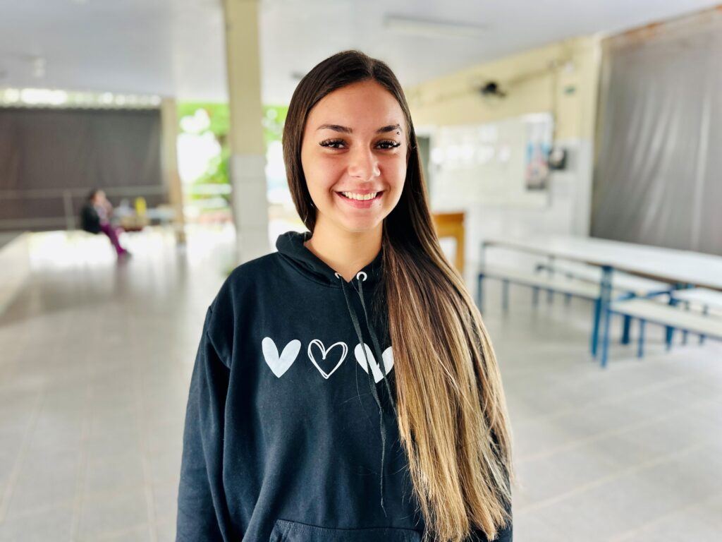 A imagem mostra uma jovem sorridente em um ambiente interno que parece ser uma escola ou refeitório. Ela está posicionada no centro da foto, com foco nítido em seu rosto, enquanto o fundo está desfocado, criando um efeito de profundidade. A jovem tem cabelos longos e lisos, castanhos com mechas loiras, e usa um moletom preto com três corações brancos estampados no peito. Ela exibe uma expressão amigável e alegre, com um pequeno piercing na sobrancelha esquerda. Ao fundo, é possível ver mesas e cadeiras, além de outra pessoa desfocada sentada, sugerindo um ambiente de convivência estudantil. A iluminação é suave e natural, destacando os tons quentes da pele da jovem.