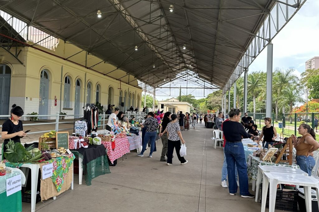 A imagem mostra uma feira ao ar livre, coberta por uma estrutura metálica com telhado, situada ao lado de um prédio de fachada amarela com janelas arqueadas. A feira é formada por várias barracas organizadas em fila ao longo de um corredor amplo, onde vendedores expõem seus produtos sobre mesas cobertas por toalhas coloridas e estampadas. Os produtos vendidos parecem ser variados, incluindo frutas, verduras, roupas e artesanatos. Há muitas pessoas circulando pelo local, algumas observando as mercadorias, outras conversando com os vendedores. O ambiente é movimentado e transmite uma sensação de comunidade e interação social. Ao fundo, é possível ver algumas árvores e um edifício mais alto, indicando que a feira está em uma área urbana. A iluminação natural é abundante, sugerindo que a foto foi tirada durante o dia. Textos visíveis em placas indicam nomes de grupos ou coletivos, como "LA HUERTITA", "COLETIVO REDE GULANDU" e "COLETIVO QUEBRADA CRIATIVA", o que reforça a ideia de que a feira pode ser organizada por iniciativas comunitárias ou grupos locais. No geral, a imagem retrata um espaço vibrante e acolhedor, onde pessoas se reúnem para comprar produtos frescos e artesanais, promovendo a cultura local e o comércio solidário.