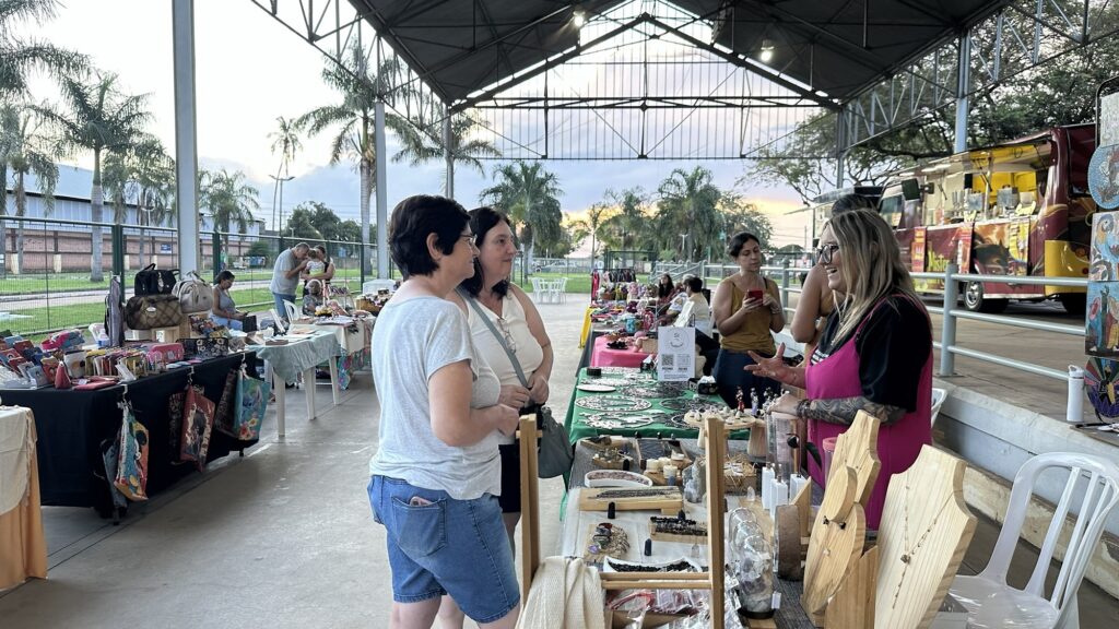 A imagem mostra uma cena de mercado ao ar livre, coberto por uma estrutura que protege as barracas do sol. O ambiente tem uma iluminação suave, possivelmente do início da manhã ou fim da tarde. No primeiro plano, há três mulheres conversando e observando os produtos expostos: uma delas está atrás da barraca, usando um colete rosa e com tatuagens nos braços, enquanto as outras duas vestem roupas casuais, uma com camiseta cinza e shorts jeans, e a outra com regata branca. A barraca principal expõe vários acessórios artesanais, como colares pendurados em suportes de madeira, brincos e outros itens decorativos. Ao lado, há outra barraca com bolsas coloridas e carteiras, algumas com estampas de personagens de desenho animado. Ao fundo, mais barracas e um food truck amarelo e vermelho são visíveis, além de árvores e vegetação, sugerindo que o mercado está instalado em uma área de parque ou lazer. O chão é de concreto e há cadeiras plásticas brancas espalhadas. A atmosfera geral é descontraída e típica de feiras comunitárias ou eventos locais.
