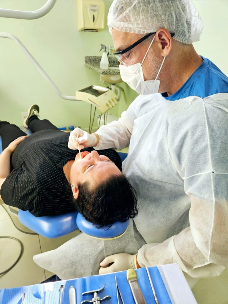 A imagem mostra um cenário típico de um consultório odontológico, onde um dentista está realizando um exame ou procedimento em um paciente. O dentista está vestido com roupas de proteção, incluindo touca, máscara, óculos de proteção, jaleco branco descartável sobre um uniforme azul, e luvas. Ele está usando um bastão de madeira, possivelmente um aplicador ou um espátula, próximo à boca do paciente. O paciente está reclinado em uma cadeira odontológica azul, vestindo uma blusa preta listrada e calça escura, com tênis verde claro. Atrás do paciente, há uma parede verde clara e uma pia com dispensadores de sabão e álcool em gel, além de uma garrafa de spray. Ao lado, há uma bandeja com instrumentos odontológicos sobre uma superfície azul, sugerindo que um procedimento ou exame detalhado está em andamento. A iluminação é clara e direcionada, típica de ambientes médicos, e a imagem foi capturada de um ângulo superior, oferecendo uma visão ampla da interação entre dentista e paciente.