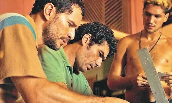 A imagem mostra um close de três homens, todos concentrados em algo fora do quadro, possivelmente trabalhando juntos em uma tarefa. O homem à esquerda veste uma camisa listrada com tons de laranja, azul e branco, tem cabelo encaracolado e barba, e está olhando para baixo com expressão séria. O homem no centro, de perfil, também tem cabelo encaracolado e veste uma camisa polo verde, igualmente focado no que está à sua frente. O homem à direita está sem camisa, com cabelo loiro estilizado e usa um colar. Ele segura um objeto longo e plano, talvez uma ferramenta ou peça de trabalho, e também está atento à atividade. O fundo é composto por ripas de madeira, sugerindo um ambiente rústico ou de oficina. A iluminação é quente, realçando os tons de pele e criando uma atmosfera de concentração e colaboração.