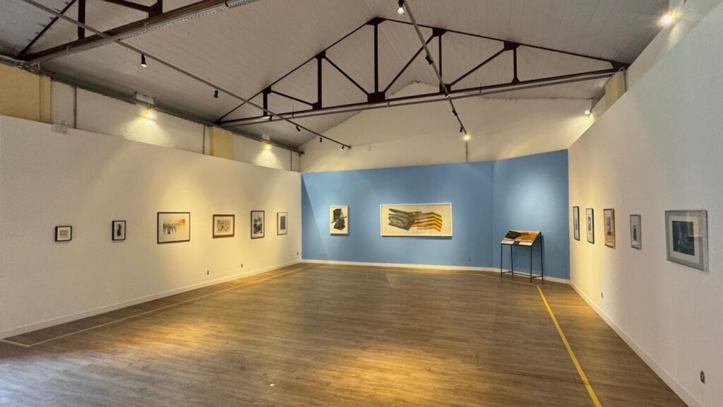 A imagem mostra o interior de uma galeria de arte contemporânea. O espaço expositivo é amplo, com paredes predominantemente brancas e uma parede destacada em azul claro, que serve como fundo para algumas obras. O chão é de madeira clara e polida, refletindo a luz. A iluminação é feita por luzes direcionadas de trilhos no teto, que destacam as obras expostas. Na parede à esquerda, há cinco pequenas obras emolduradas, provavelmente fotografias ou gravuras. Na parede azul, duas obras maiores chamam a atenção: uma escultura ou pintura abstrata escura e uma pintura abstrata horizontal, colorida, com formas geométricas em tons de amarelo, laranja e verde. À direita, há um suporte metálico preto com livros ou portfólios de arte e mais obras emolduradas, incluindo uma peça maior no final da parede. O teto é industrial, com vigas metálicas expostas. O ambiente está vazio, sem pessoas, e o chão tem fitas amarelas que provavelmente indicam limites ou caminhos para os visitantes. A imagem transmite uma atmosfera clara, organizada e convidativa para a apreciação das obras expostas.