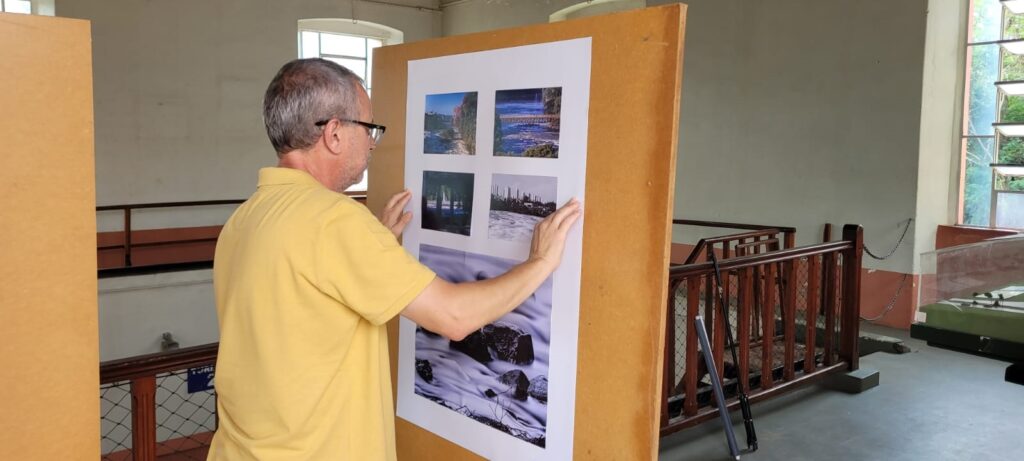 A imagem mostra um homem de meia-idade, vestindo uma camisa polo amarela, que está de perfil enquanto ajusta ou fixa um grande cartaz branco em um painel de madeira marrom. O cartaz contém cinco fotografias: quatro pequenas no topo alinhadas horizontalmente e uma maior na parte inferior. O ambiente parece ser uma galeria ou museu com paredes brancas, teto alto e janelas grandes que permitem a entrada de luz natural. Ao fundo, há uma grade de madeira com tela metálica, sugerindo que o local pode ser um espaço industrial adaptado para exposições. A atmosfera é tranquila e o foco está no homem enquanto ele organiza a exposição.
