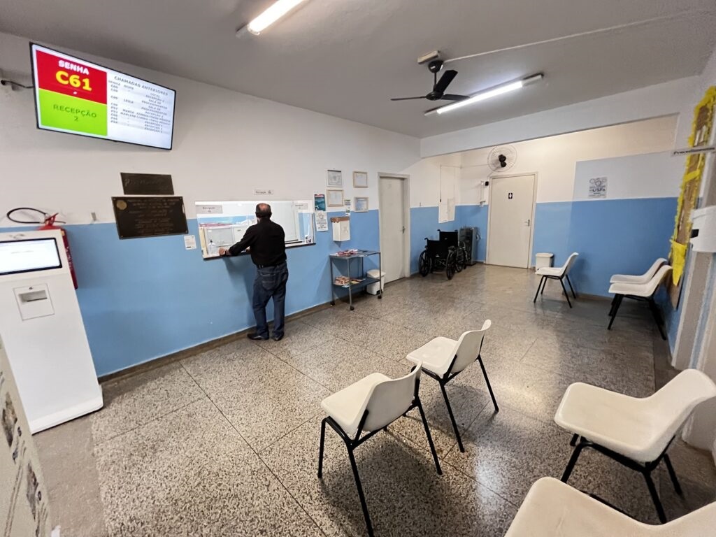 A imagem mostra uma sala de espera, provavelmente em um ambiente público como uma clínica, hospital ou repartição pública. No centro, há um balcão de atendimento com uma janela onde um homem está sendo atendido. Acima do balcão, um painel eletrônico exibe a senha atual "C61" para o setor de "Recepção 2". À esquerda do balcão, há um equipamento eletrônico, possivelmente um terminal de autoatendimento ou um quiosque para retirada de senhas. A sala tem paredes pintadas em azul claro e branco, e o piso é de um material cinza com manchas. À direita, há várias cadeiras de plástico branco com estrutura metálica preta, organizadas para os clientes aguardarem sua vez. Ao fundo, há duas cadeiras de rodas próximas a uma porta, indicando acessibilidade para pessoas com mobilidade reduzida. O teto possui luminárias fluorescentes e um ventilador de teto. No painel eletrônico, também são exibidas diversas senhas anteriores atendidas, como 148, 168, 169, entre outras, além de nomes como Maria, Mariana e Marcelo, sugerindo o sistema de chamada utilizado no local. O ambiente é simples, funcional, com foco na organização do atendimento, transmitindo uma atmosfera típica de serviços públicos ou de saúde.