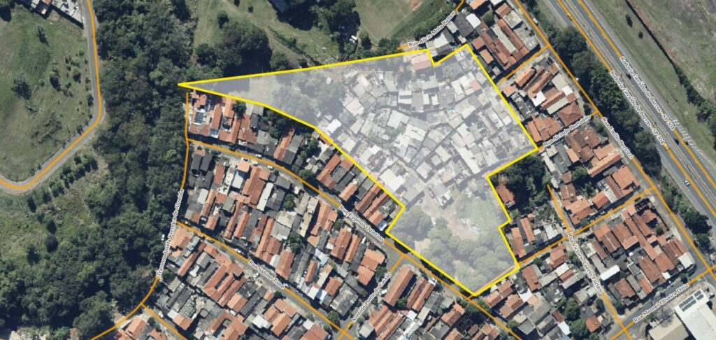 A imagem mostra uma vista aérea de uma área urbana densa, provavelmente uma favela, destacada por um contorno amarelo. As construções são muito próximas umas das outras, com telhados predominantemente vermelhos, indicando casas simples e pequenas. A favela está cercada por áreas residenciais mais organizadas e algumas áreas verdes com árvores. Ao lado da comunidade, há uma rodovia movimentada chamada Rodovia Geraldo de Barros - SP 304, que delimita uma das bordas da área. Várias ruas dentro e ao redor da favela são identificadas, como Rua Sabino Tome, Avenida Ponte do Caixão e Rua Emílio Bertozzi. A imagem evidencia o contraste entre a densidade e informalidade da favela e a infraestrutura mais estruturada da região ao redor, além da presença de áreas naturais próximas. A iluminação sugere que a foto foi tirada durante o dia, com boa visibilidade e detalhes claros das construções e ruas.