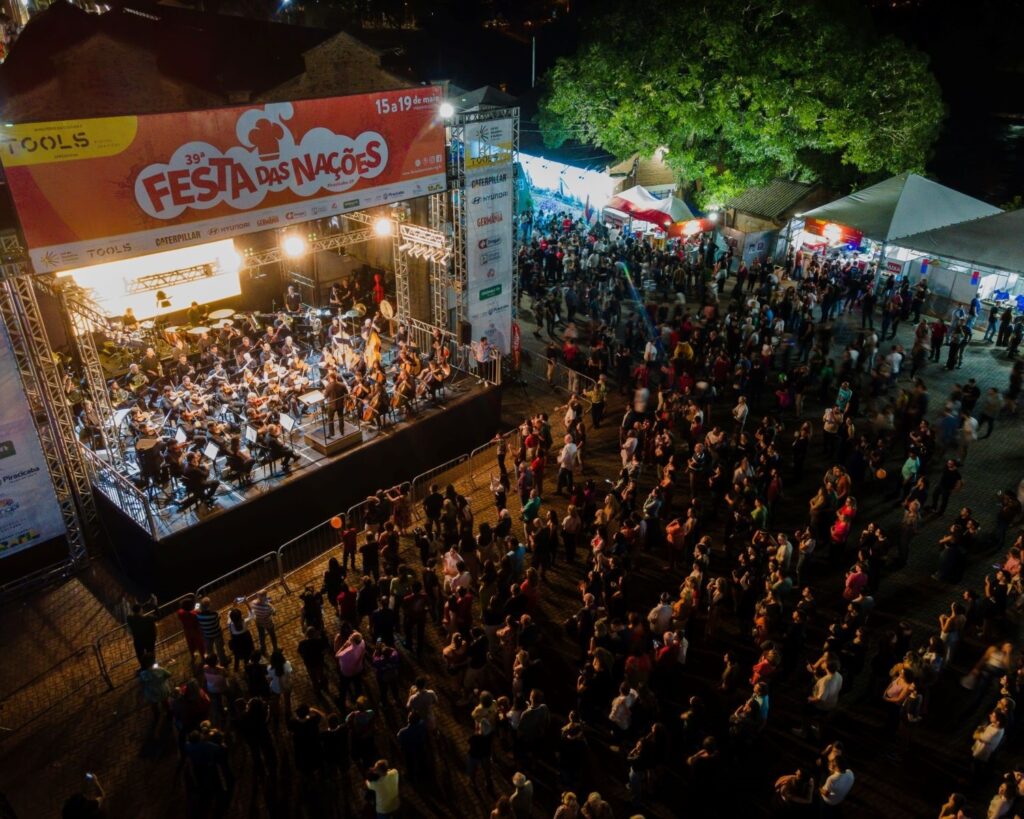 A imagem mostra uma grande festa ao ar livre à noite, chamada "39ª Festa das Nações", realizada em Piracicaba, São Paulo, entre os dias 15 e 19 de maio. No centro da cena, há um palco iluminado onde uma orquestra está se apresentando, com vários músicos tocando instrumentos como violinos, violoncelos e metais. O palco está decorado com um grande banner que exibe o nome do evento e os patrocinadores, como TOOLS, CATERPILLAR, HYUNDAI e GERMANIA. À frente do palco, uma grande multidão de pessoas assiste ao espetáculo, muitas delas segurando celulares para registrar o momento. Ao redor, é possível ver tendas e barracas que provavelmente oferecem comida, bebida ou outras atrações típicas de uma festa. À direita da imagem, há uma árvore grande que se destaca no cenário urbano. A iluminação quente do palco contrasta com o céu escuro da noite, criando um ambiente vibrante e festivo. A perspectiva aérea permite uma visão ampla do evento, mostrando a extensão do público e a organização do espaço. A imagem transmite a energia e a diversidade cultural do festival, que reúne música, público animado e uma atmosfera de celebração comunitária.