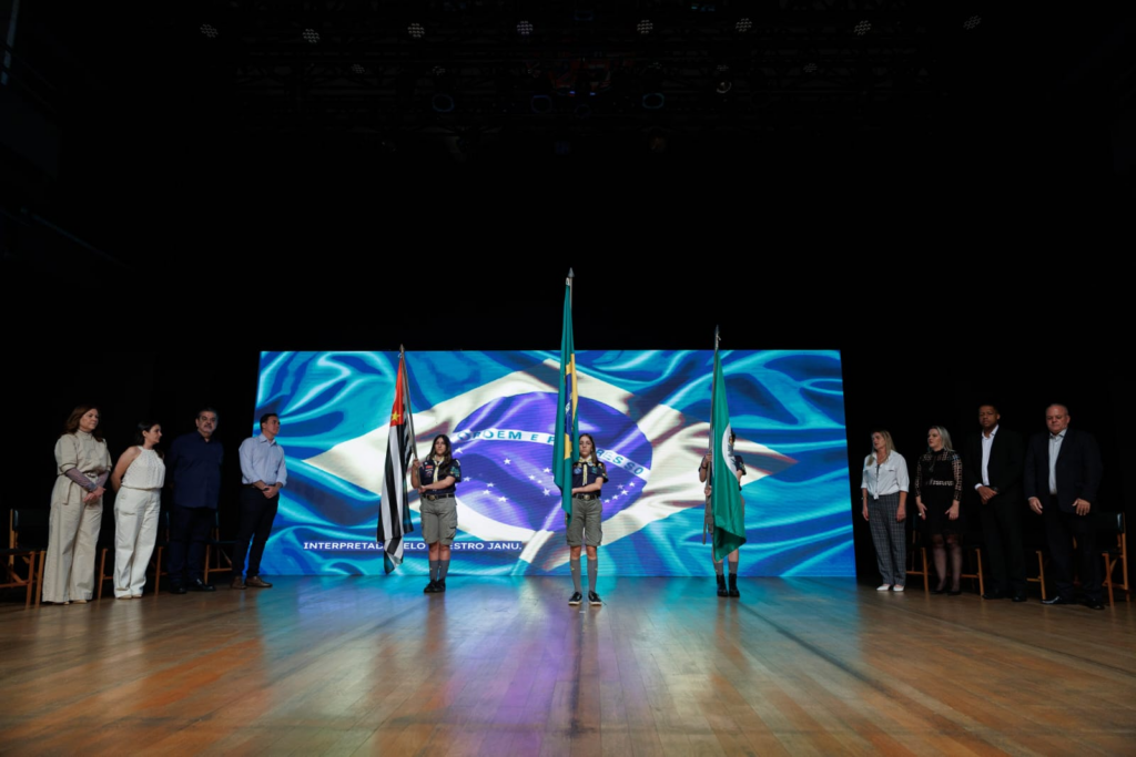 A imagem mostra uma cerimônia formal acontecendo em um palco com um grande telão ao fundo exibindo a bandeira do Brasil. No centro do palco, três jovens vestidos com uniformes de escoteiros estão em posição de atenção, segurando bandeiras — duas delas são bandeiras do Brasil e a outra parece ser uma bandeira estadual, possivelmente do estado de São Paulo, devido às listras pretas e brancas e a estrela vermelha. À esquerda dos escoteiros, cinco adultos estão em pé, observando a cerimônia, vestidos com roupas formais. À direita, há mais quatro adultos também vestidos formalmente. O ambiente transmite uma atmosfera solene e patriótica, com iluminação focada nos participantes e o palco de madeira refletindo a luz. O telão em azul e verde destaca a bandeira brasileira e cria um cenário vibrante para o evento.