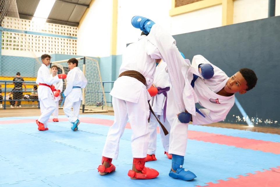 A imagem mostra uma cena vibrante de uma aula de karatê, provavelmente realizada em um ginásio coberto com grandes janelas que deixam entrar bastante luz natural. No centro da imagem, dois meninos estão em pleno combate. O garoto à direita está executando um chute alto e ágil, com o corpo no ar, usando kimono branco, protetores azuis nas mãos e nos pés, e faixa azul. Ele está concentrado e determinado. O adversário, à esquerda, também veste kimono branco, mas tem protetores vermelhos e faixa marrom, e parece estar se preparando para bloquear ou reagir ao chute. Ao fundo, outras crianças, também vestindo kimonos e protetores vermelhos e azuis, observam ou aguardam sua vez. O chão é revestido com tatame nas cores azul e vermelho, típico para práticas de artes marciais. A atmosfera transmite energia, disciplina e a dedicação dos jovens atletas ao esporte.