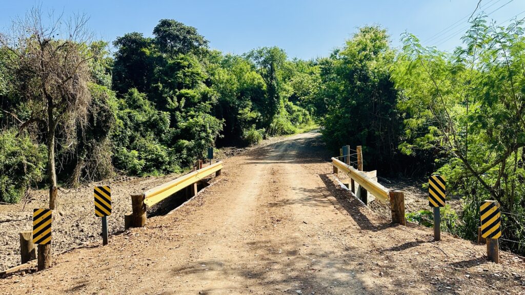 A imagem mostra uma estrada de terra que atravessa uma pequena ponte. A estrada é cercada por vegetação verde e árvores, algumas delas com folhas secas, contrastando com as outras mais verdes. O céu está claro e azul, indicando um dia ensolarado. A ponte possui guardrails amarelos e postes de madeira, além de placas de advertência com listras amarelas e pretas em ambos os lados da entrada. A superfície da estrada é irregular, com marcas de pneus visíveis. A vegetação ao redor é densa e variada, criando um cenário natural e tranquilo. A iluminação forte, típica do meio-dia, destaca as texturas do solo e das folhas. Não há pessoas ou animais na cena, e a perspectiva da foto é baixa, olhando para cima em direção à ponte e à estrada que segue adiante.